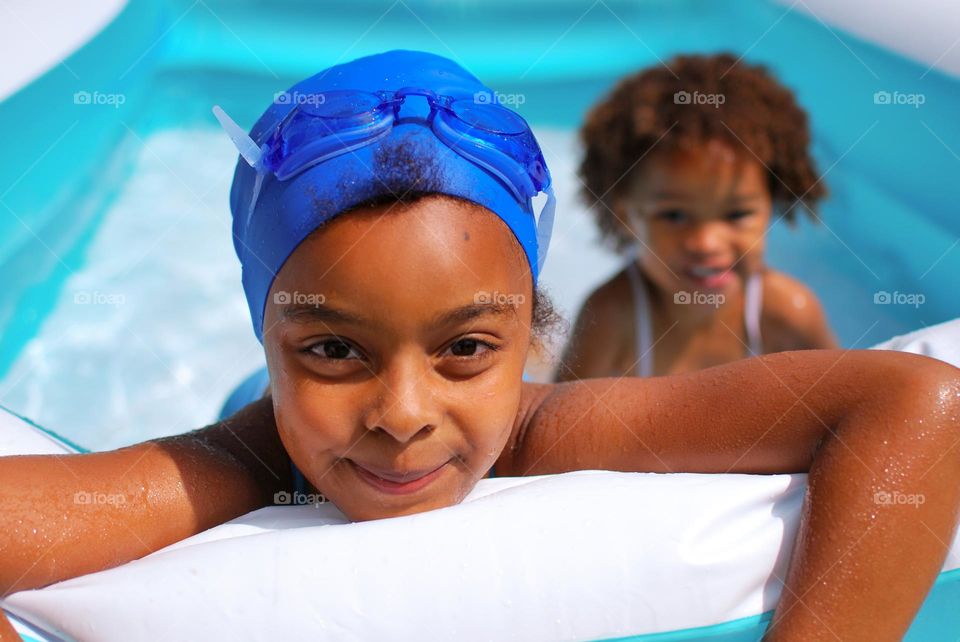 Girl of mixed race enjoying the refreshment of water in a swimming pool on a hot summer day, together with her little sister (family, fun, summer, water, blue, swimming suit, splash, hot, enjoy, play, outdoors)