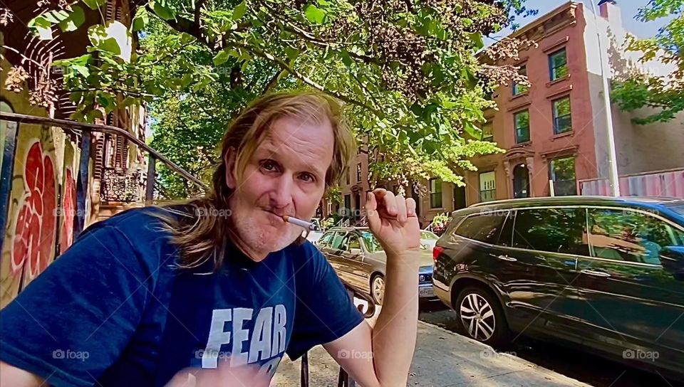 My partner for nearly 16 years, “Timothy Trespas” is enjoying his cigarette sitting on our favorite stoop on “Waverly St” in “Clinton Hill”, Brooklyn. This residential city block has charme and lush green vegetation. 2022. Hypnotic Productions