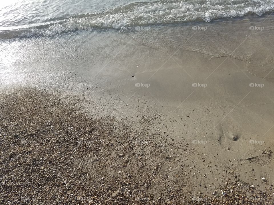 A sandy seashore showing some ocean water but mainly the sand and pebbles.