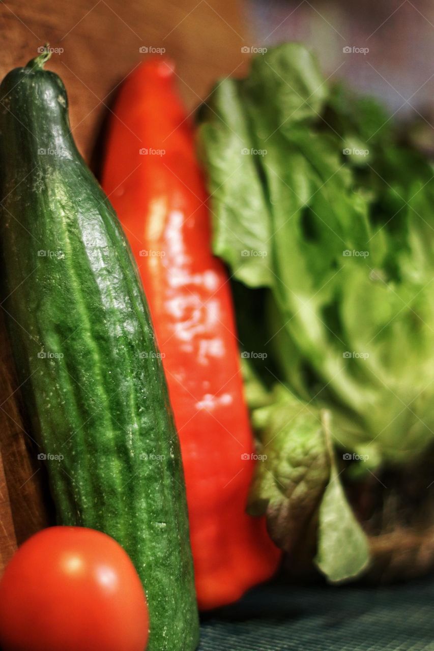 A green cucumber stands next to a tomato, a red bell pepper and lettuce on a cutting board 