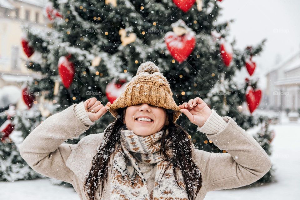 Happy young woman pulling knit hat down over her eyes, standing outdoor in front of christmas tree on a snowy day in winter