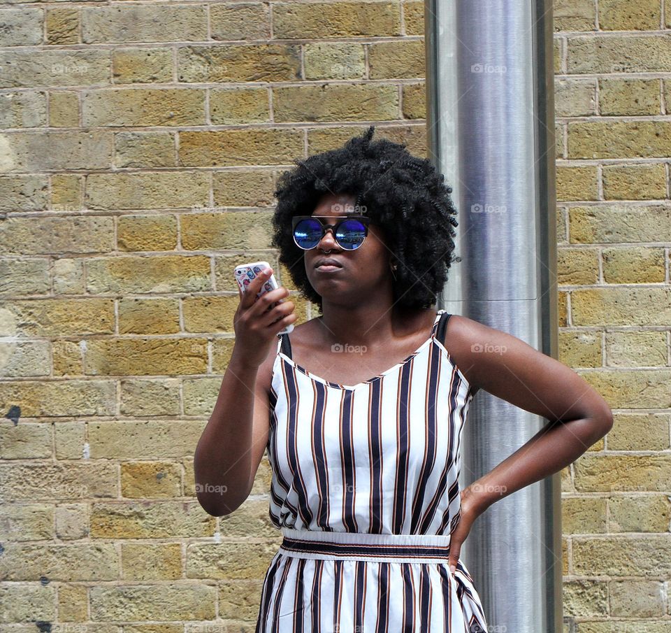 A young black girl with a large Afro hairstyle and designer, blue sunglasses looking at her mobile phone on the city streets.