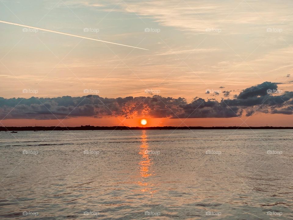 Sunset In The Horizon With Deep Orange Color Sighting  By The Island Reflecting On The Water, Under The Long Clouds.