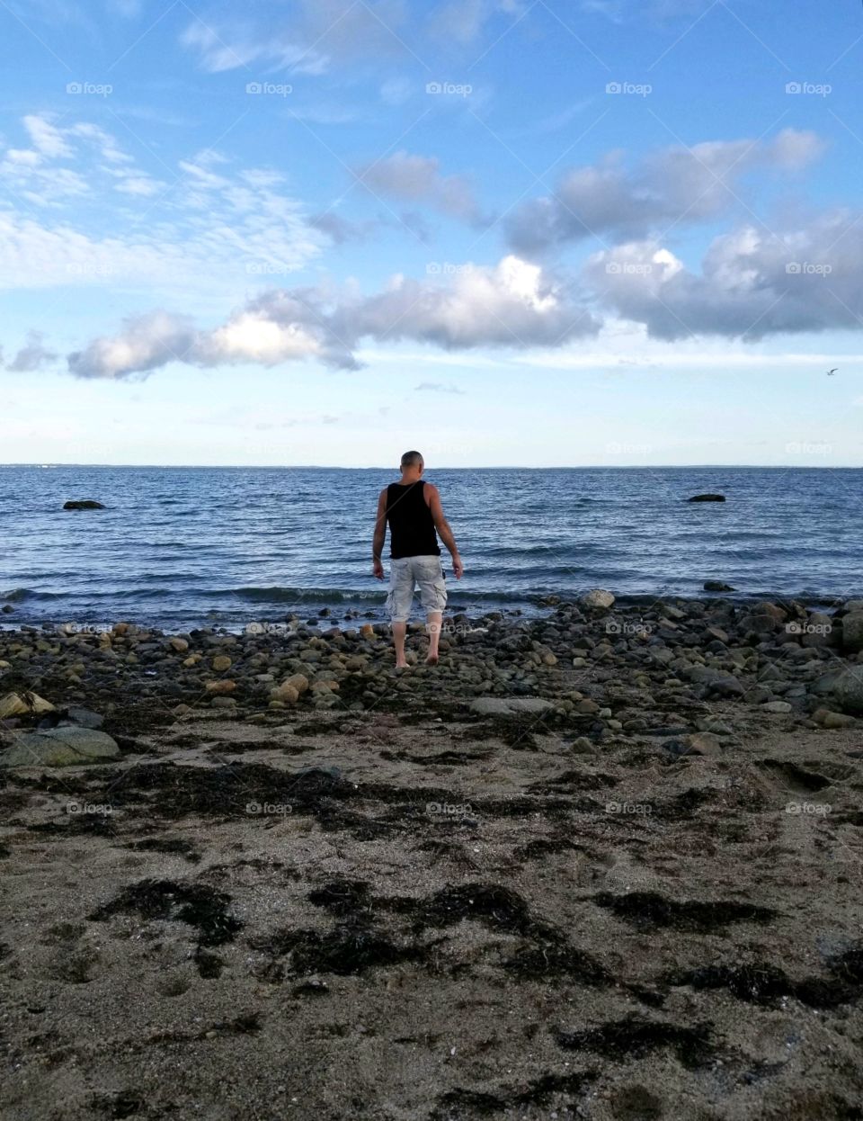Man walking on rocky beach barefoot, towards the ocean water. It's near sunset showing cloud formations.