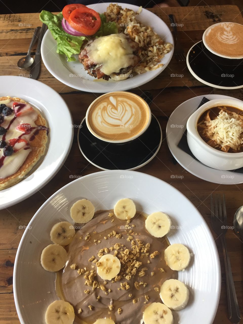Photo of brunch for two- açaí bowl with bananas, granola, and honey with a lavender latte, a chai latte, a burger with soup and a berry covered pancake 