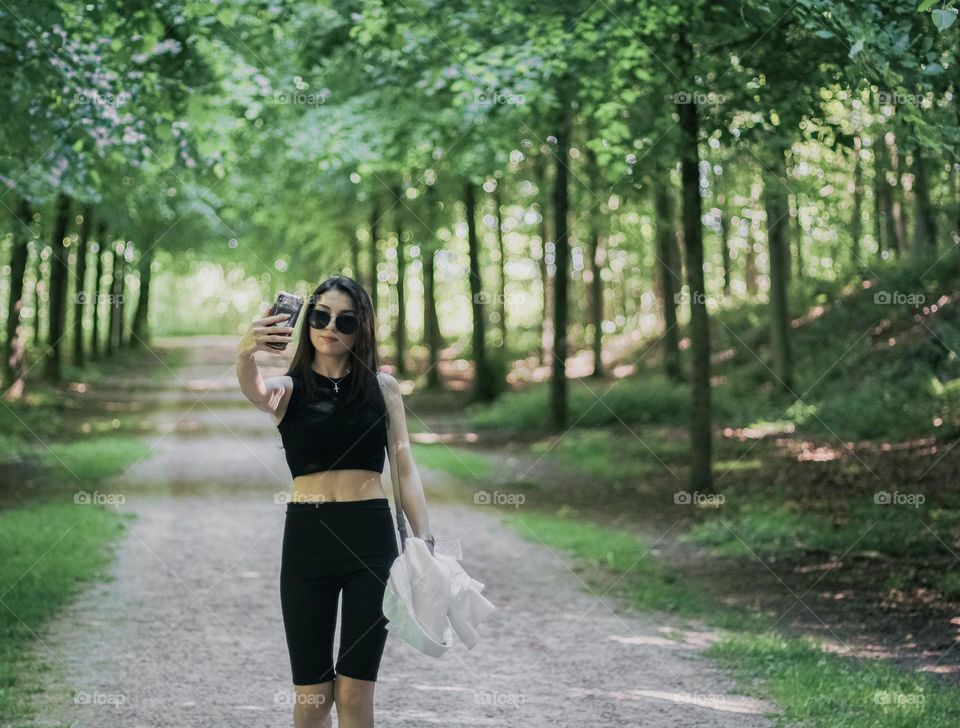 Portrait of a beautiful young caucasian brunette girl with long hair,sunglasses, in black sportswear makes a selfie with a smartphone,holding a white shirt in her hand while walking through the forest on a clear sunny day,close-up side view .Using te