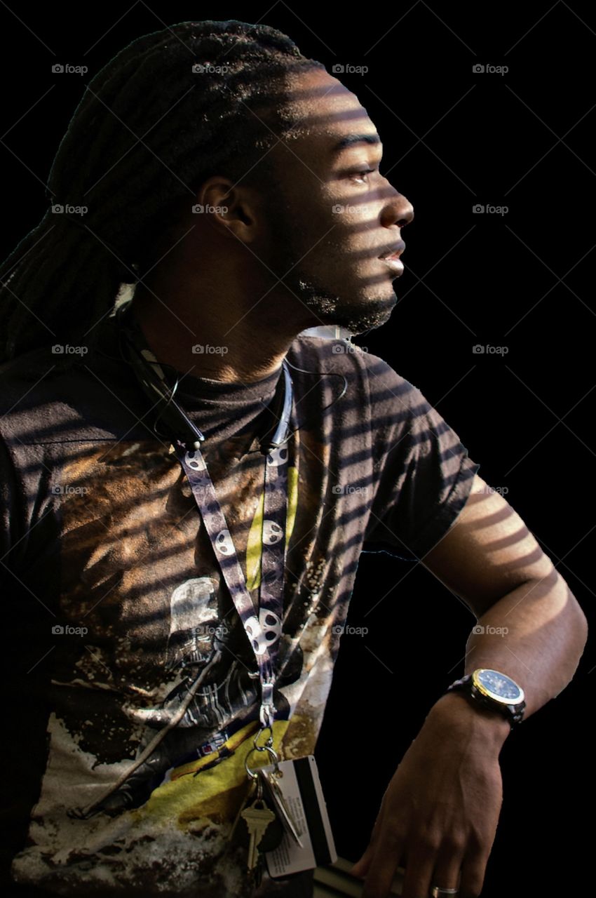 Young man looking out the window and the shadow of the blind is on his face and body 