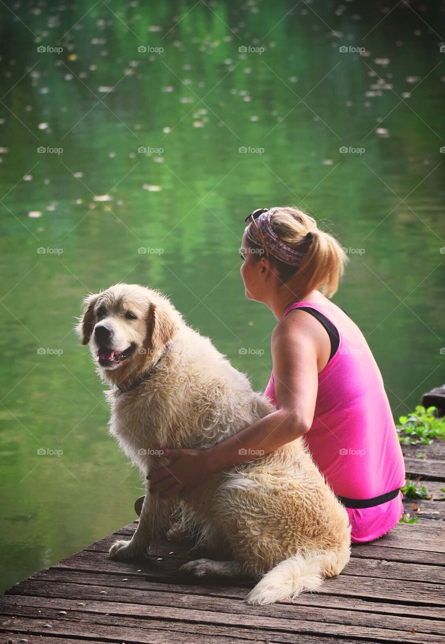 Beautiful young woman siting at the dock of a lake with her dog, a golden retriever who took a bath in the lake. Quality time on a bright and sunny spring day.