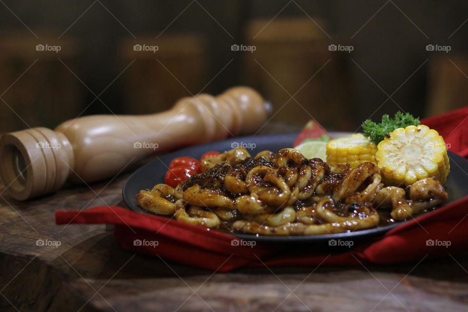 Close-up portrait of squid rings seasoned with barbecue sauce served on a black plate on a wooden table, wooden barbecue spice shaker background — Stock Photo