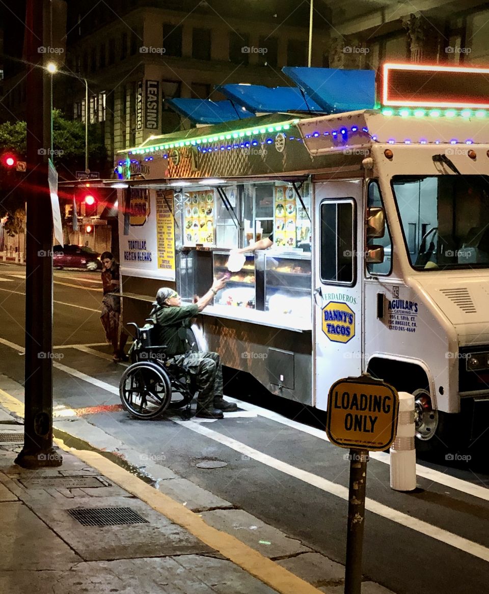 Man in a wheelchair reaches for a package being handed down from a Taco Truck parked in a Loading Zone on 7th Street in Downtown Los Angeles CA. 6.10.2020