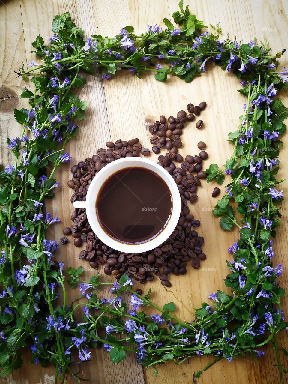 Composition.  White cup with black coffee, roasted coffee beans around.  Around a cup with coffee and beans  a frame of blue wildflowers