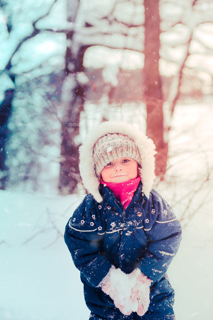 Happy smiling little girl enjoying snow. Toddler is playing outdoors in wintertime while snow falling. Toddler is wearing dark blue snowsuit and wool cap