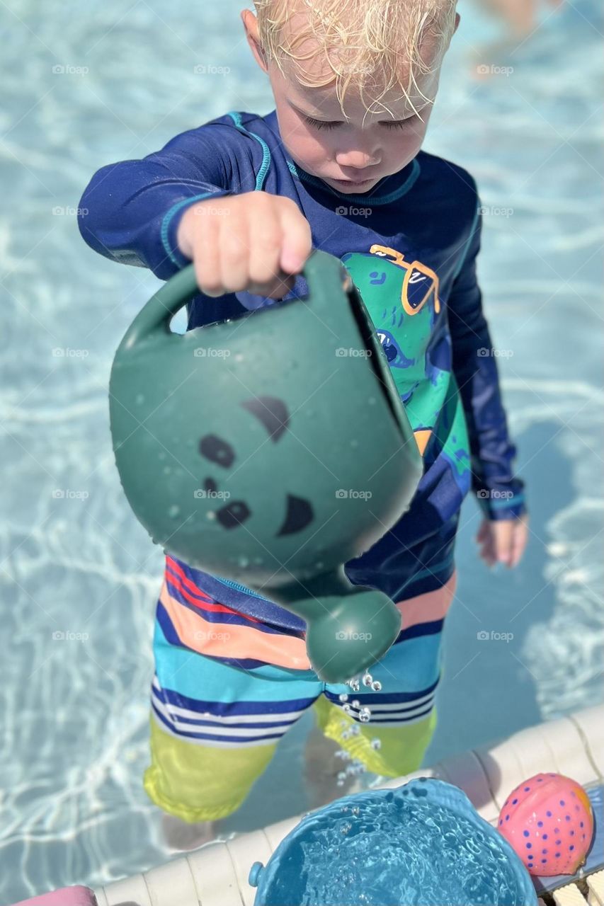 A young boy plays with toys in a pool on a sunny summer day, experimenting with the water in the shallow side
