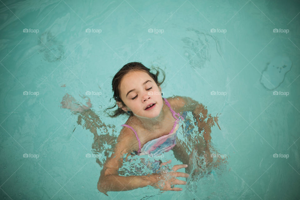 Little, happy, brunette girl floating in the pool with her eyes closed and a smile on her face. 