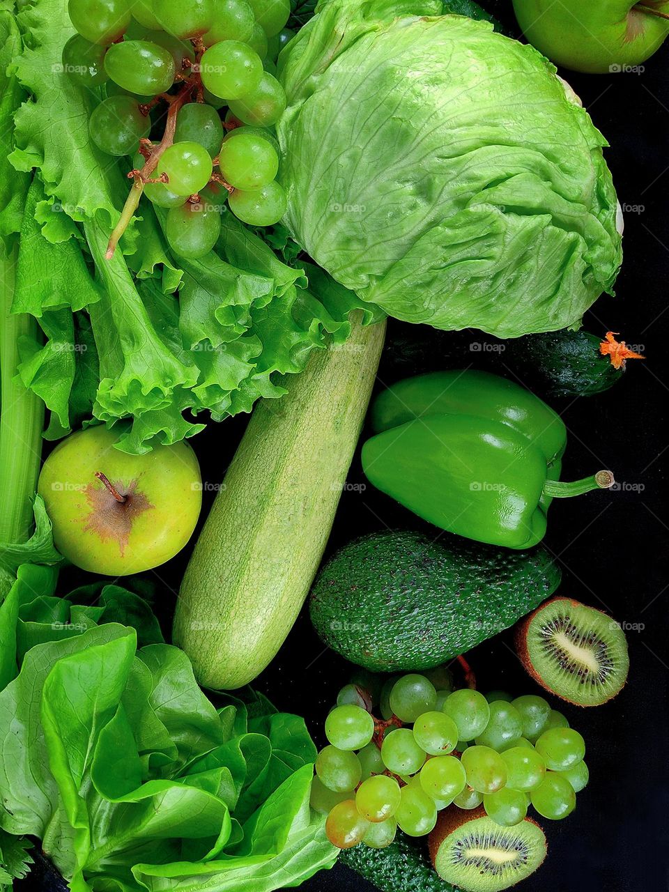 Green color. Green fruits and vegetables on a black background. Celery stalk, green apples, cucumbers, avocado, zucchini, two bunches of grapes, lettuce, iceberg lettuce, lettuce latuc, bell pepper and kiwi halves