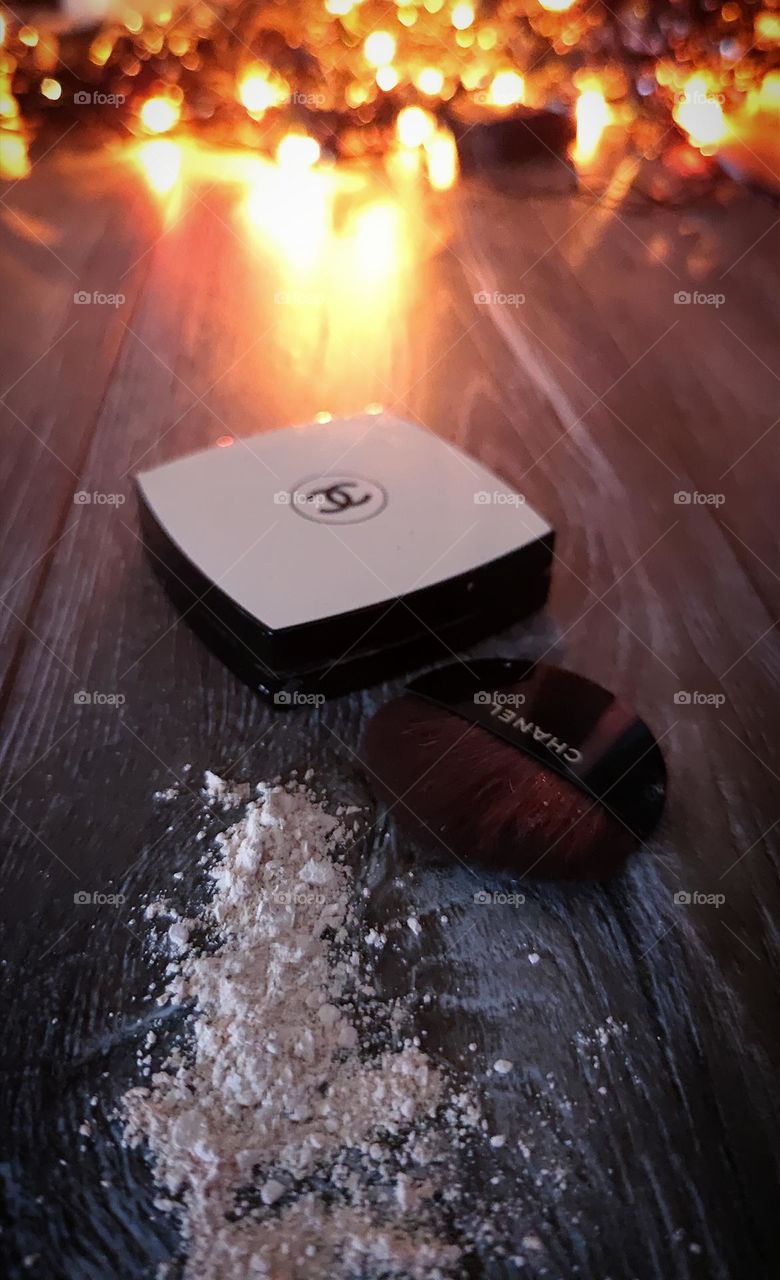 A garland is lit on a wooden background, the light from which falls on a box with the Chanel logo, on powder and a brush with the inscription "CHANEL". Powder Chanel Les Beiges