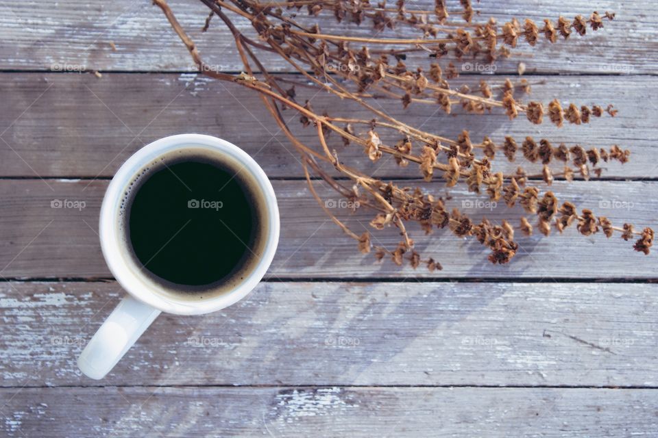 Flat lay of coffee in a white mug next to a dried brown plant on a weathered wooden surface