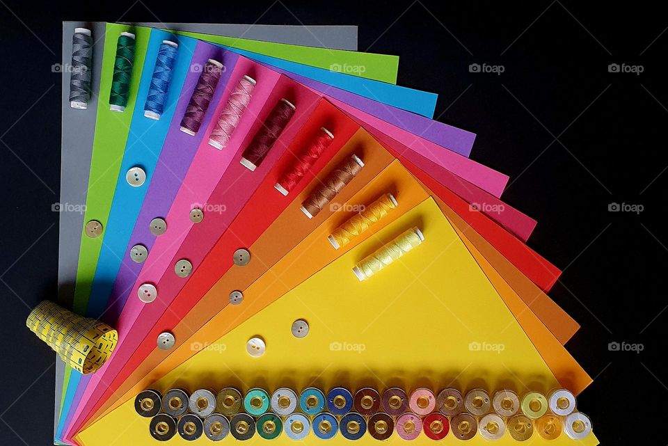 Primary colours flatlay on a black surface composed of a fan-shaped set of colour papers, sewing thread rolls, sewing machine bobbins, mother-of-pearl buttons and measuring tape