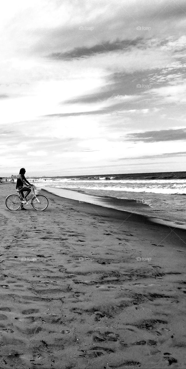 Black and white image of a woman at the beach on a bicycle in the sand while looking out at the ocean 