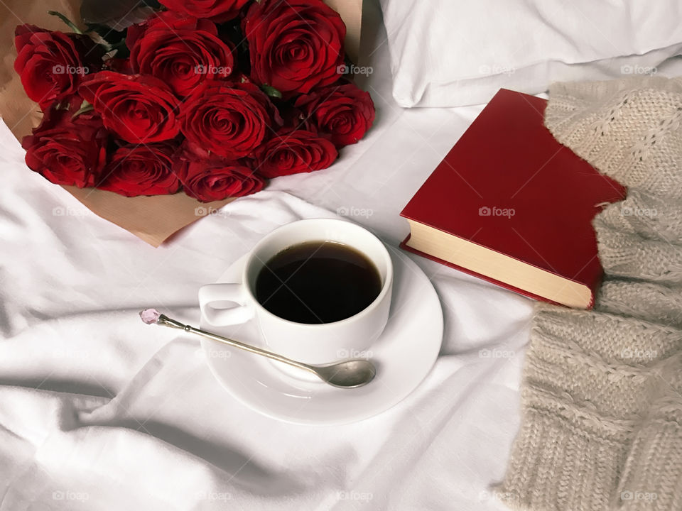 White cup with black coffee in white bed with red roses and red book