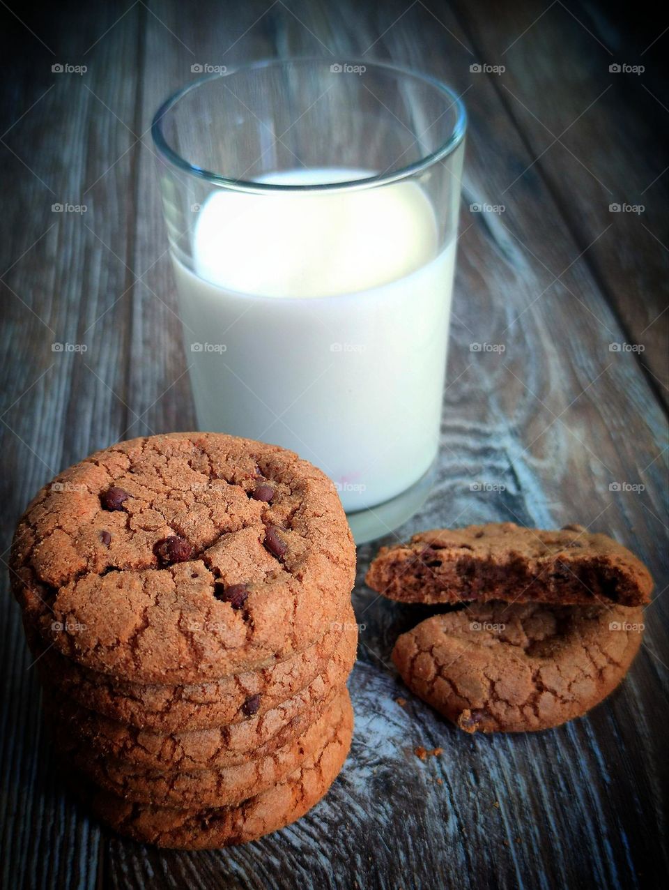 Chocolate chip cookies stacked on top of each other. Nearby is a broken chocolate chip cookie. In the background is a glass of milk. Wooden background
