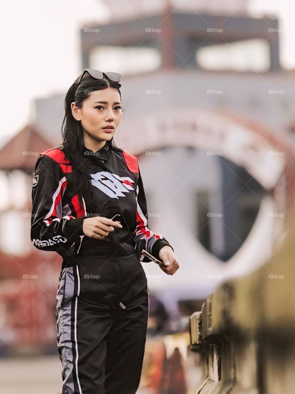 The young woman wears a stylish racing suit, predominantly black with red and white accents. Sunglasses rest on top of the head