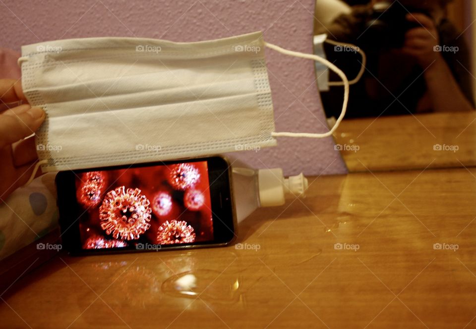 In the photograph there is a mask, disinfectant gel and an iPhone. The iphone has the image of a virus. Behind there is also a mirror where the camera comes out.