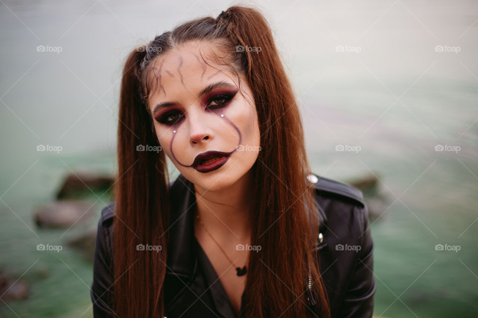 Cheerful scary woman with Halloween makeup with pale face wears costume for carnival party, posing against the backdrop of water, having fun at the holiday. A creative idea for a masquerade.	