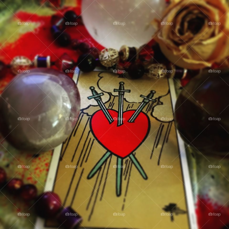 The three of swords in the Rider Waite Smith tarot deck displays three swords piercing a vulnerable heart. The card represents heartache and loss, insecurity and heartbreak. Posed with dried roses and amethyst.