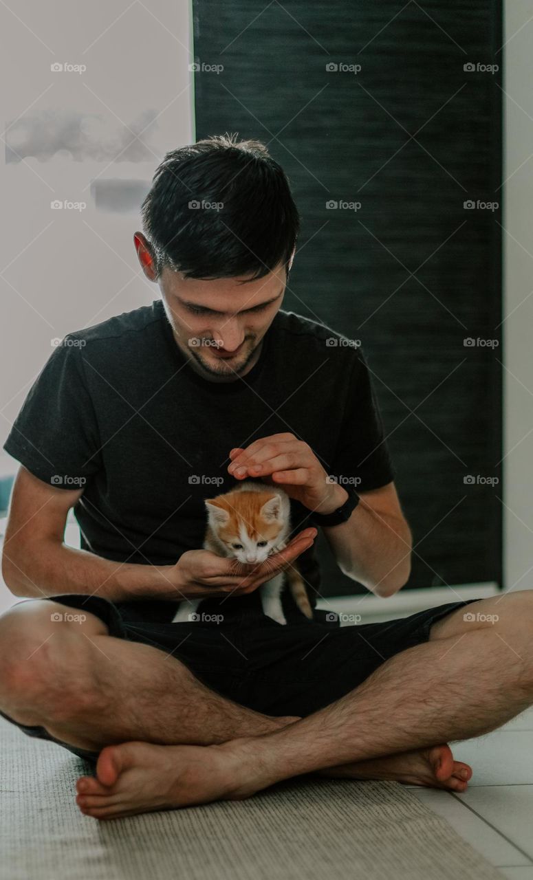 Portrait of one young handsome Caucasian guy carefully holding a small kitten in his hands and gently stroking it while standing on the floor with his legs crossed, from the side, close-up.