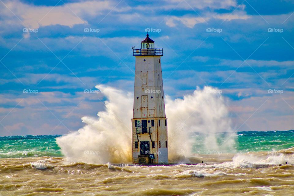 Waves splashing and crashing into a lighthouse with the bright blue sky background.