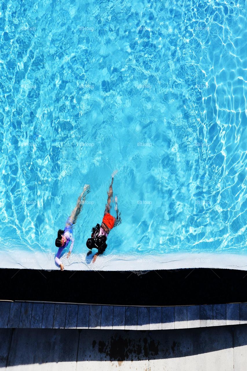 Two sisters on vacation enjoy a hot days worth of swimming in an emerald blue pool.