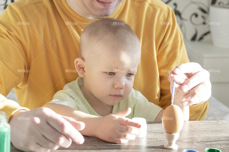 A little son and his father together in their free time paint Easter, chicken eggs with paints and brushes for the holiday. A child and a man create a craft and natural product in their family business.