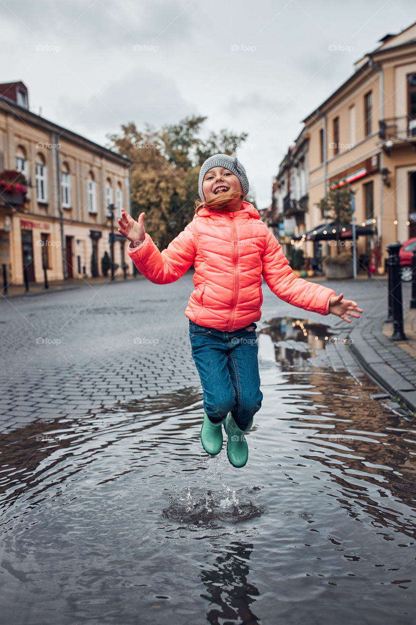 Happy smiling girl jumping in the puddle during walk in a downtown on rainy gloomy autumn day