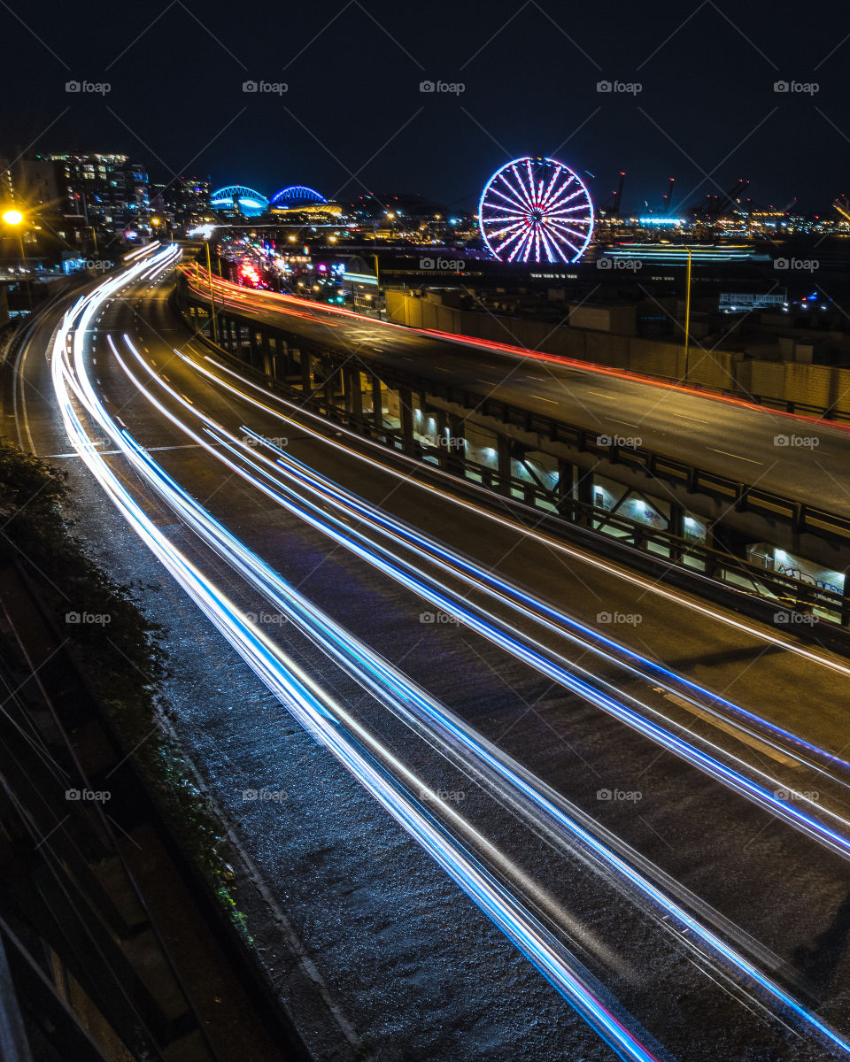 Light trails leading down a highway towards downtown Seattle, with the Seattle Wheel lit up in the top right third. 