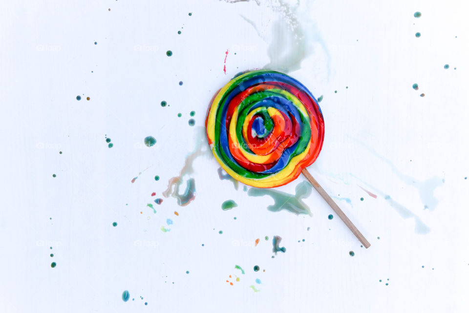 Flat lay of a large rainbow lollipop surrounded by colorful sugar splatter on a white backdrop