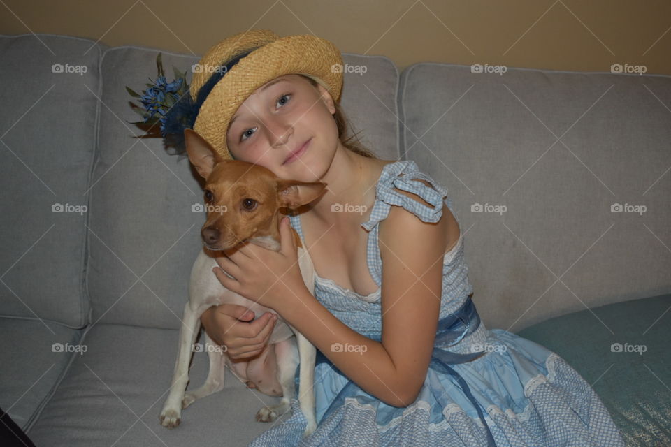 A pretty young girl in a hand made blue dress and complementing straw hat, hugs her rat terrier dog, sitting on a sofa. 