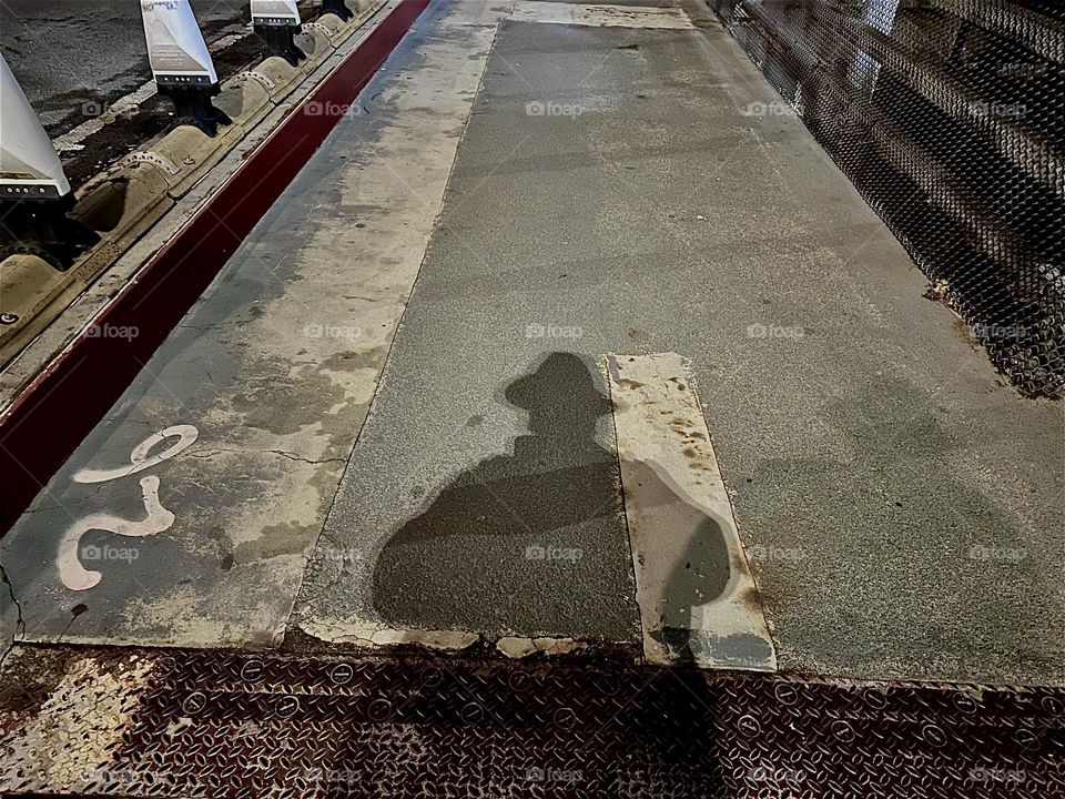 A dramatic shadow is cast onto the pedestrian lane of the “Pulaski Bridge” at “Newtown Creek”, the bridge that connects “Greenpoint”, Bklyn and LIC, Queens. The nighttime illumination brings out the most compelling effects. 2023. Hypnotic Productions