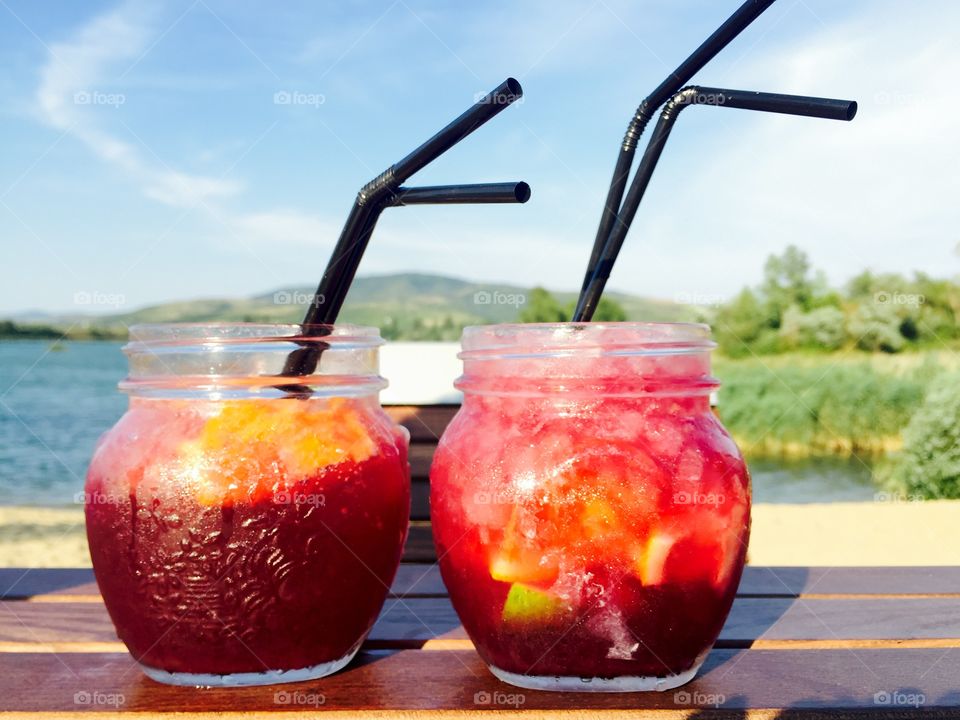 Two colourful glasses with strawberry lemonade on wooden table with lake in the background 