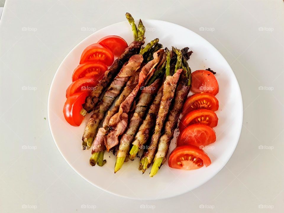 Close up of the white plate with tomato slices and green asparagus wrapped in bacon