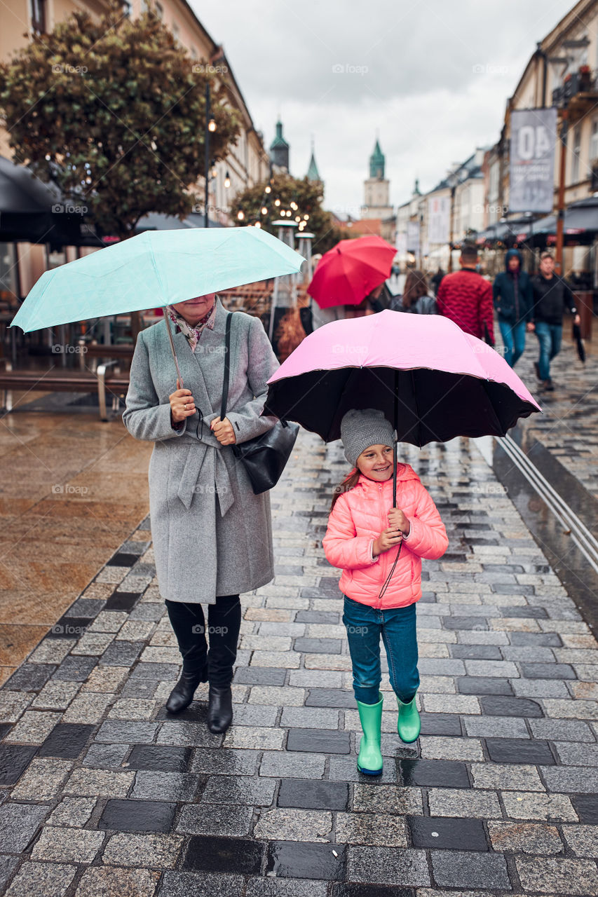 Mother and her little daughter holding the pink and blue umbrellas walking in a downtown on rainy gloomy autumn day