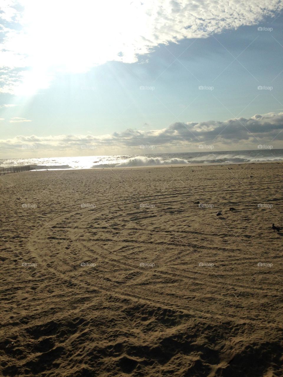 A sandy beach with circular tracks in it underneath a partly cloudy September sky. The whitecaps in the water resemble the clouds above, and a bit of sun peaks through and reflects on the water. Taken in Point Pleasant Beach, NJ.