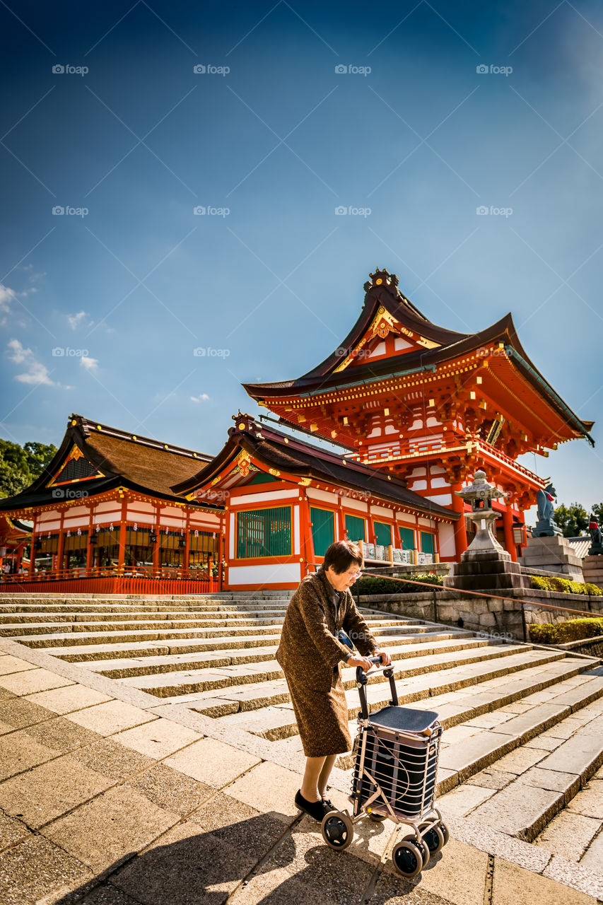 Elderly lady taking a morning stroll around temple grounds. Aided by a trolley or cart, she walked slowly down a slope. At the Fushimi Inari Taisha Shrine, Kyoto, Japan.