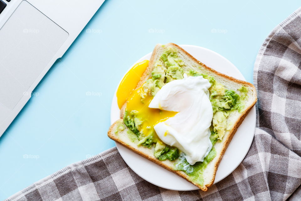 Sandwich with fresh green avocado, poached egg on white plate lying on blue background 