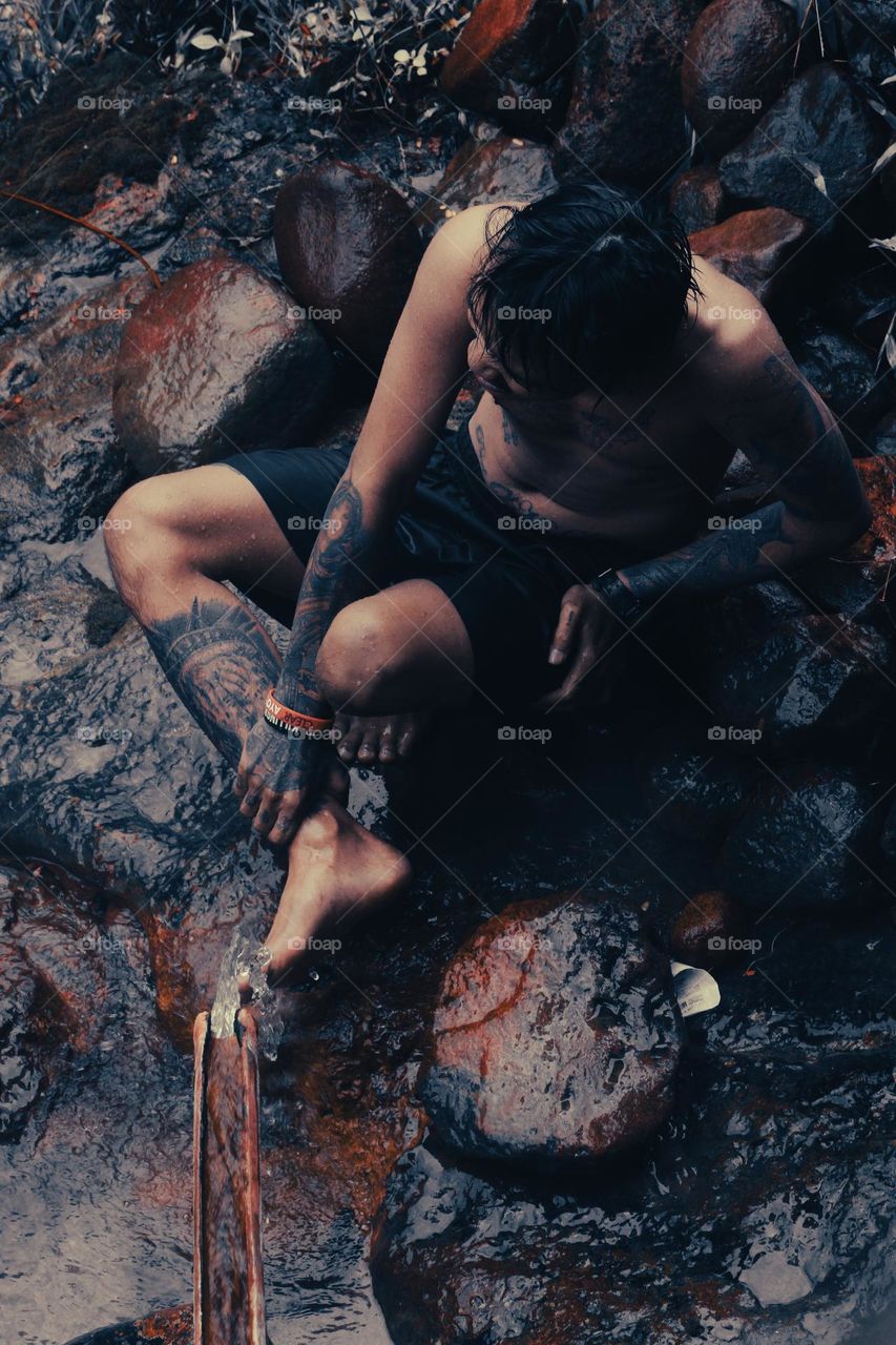 Under a bamboo shower, a tattooed man sits on a mossy rock holding his right leg, both hands and feet covered in tattoos.