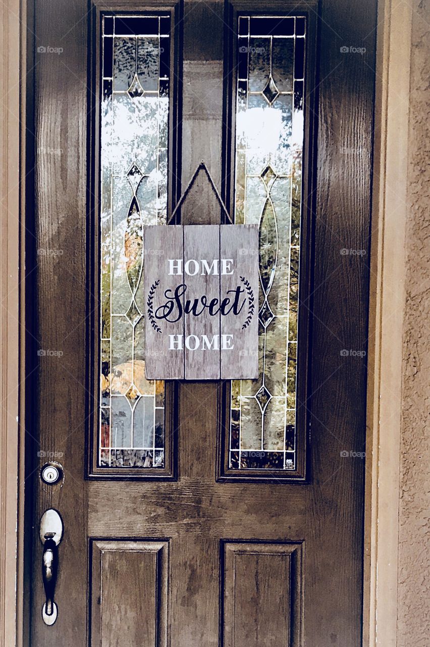 A wooden stained front door with a wooden sign saying home sweet home displayed against the glass window. 