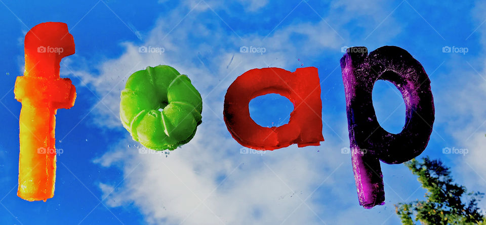 Clash of Colours: Orange, green, red & purple frozen gelatin letters spelling FOAP. The letters are sitting on a mirror which is reflecting a bright blue sky dappled with clouds & an overhanging branch of a nearby tree. 