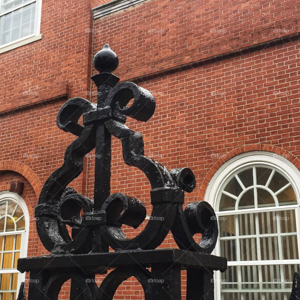 Decorative iron work sits atop a wrought iron fence with the backdrop of a classic brick building.
