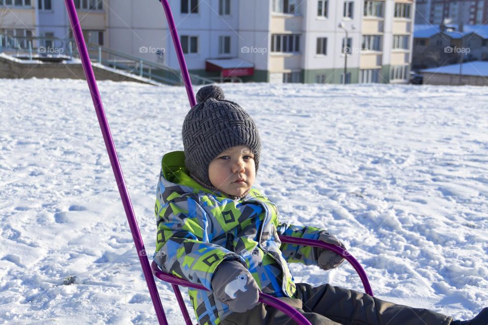 A child in winter in winter jackets, pants, a hat and boots on white snow on the street and in the park in nature plays winter fun and sleds.