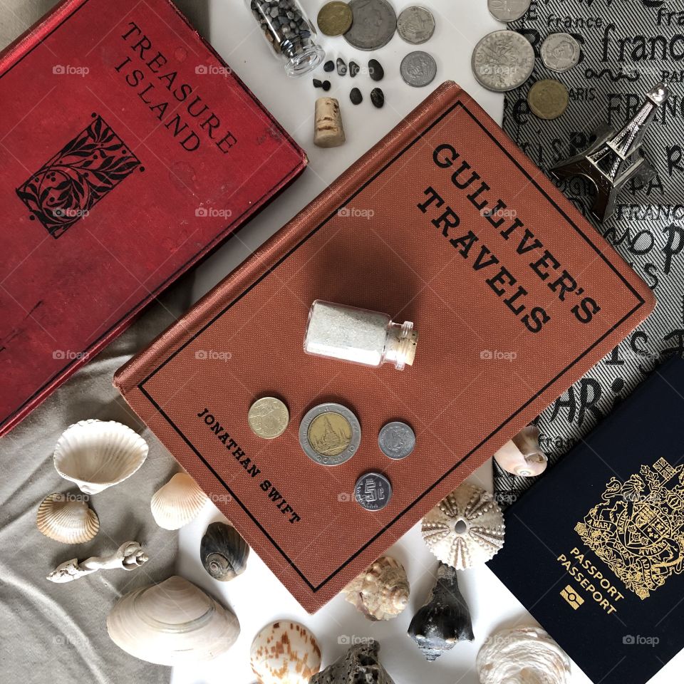 Hey vintage copy of Gullivers travels is surrounded by articles of travel and leisure such as a passport, foreign currency, shells, sand, and a small Eiffel Tower